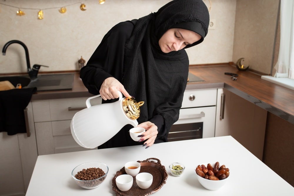 young woman in black hijab pours arabic coffee from a white dallah into a small cup on a decorated tray, with dates and cardamom nearby, showing a traditional hospitality moment at home
