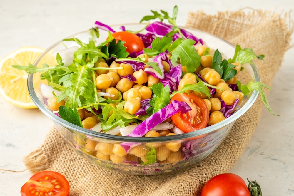 Salad with chickpeas, arugula, tomatoes, red cabbage and herbs in a bowl on a cloth on a light background.