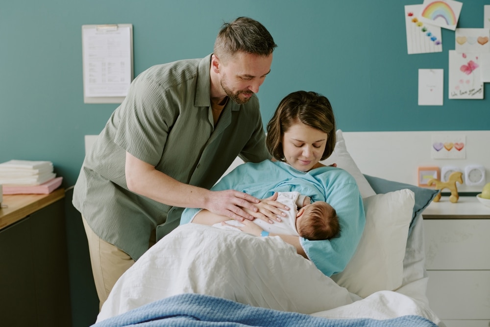 Young couple tenderly caring for their newborn baby in comfortable hospital room. Father standing beside bed, mother lovingly holding and feeding baby