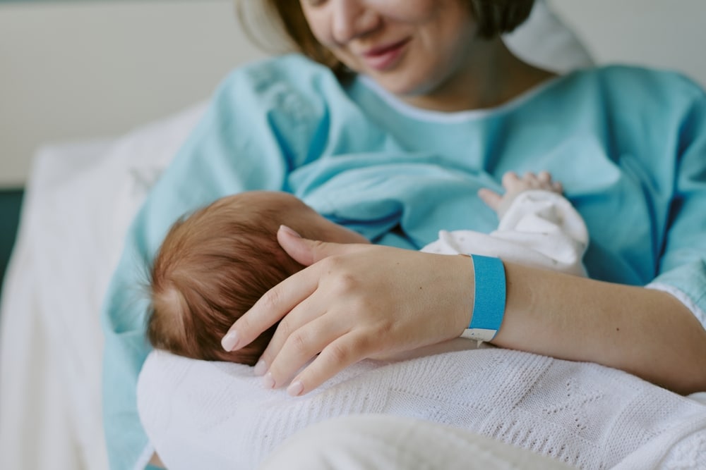 New mother in hospital gown breastfeeding newborn baby while holding closely. Both mother and baby are sharing a tender bonding moment