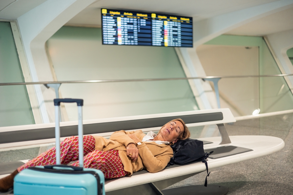 Senior woman sleeping on a bench at the airport terminal while waiting for her flight departure, next to her light blue trolley and backpack