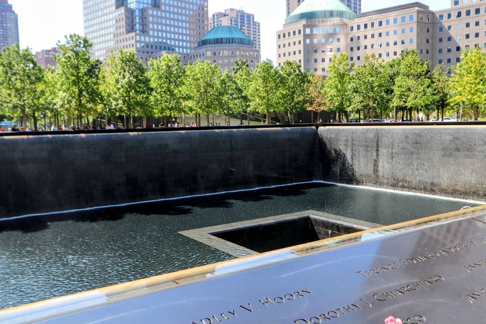 NYC, NY - Sept 13, 2025 - A view of the National September 11 Memorial Museum in New York City, featuring one of the twin reflecting pools surrounded by trees and a row of skyscrapers in a sunny sky