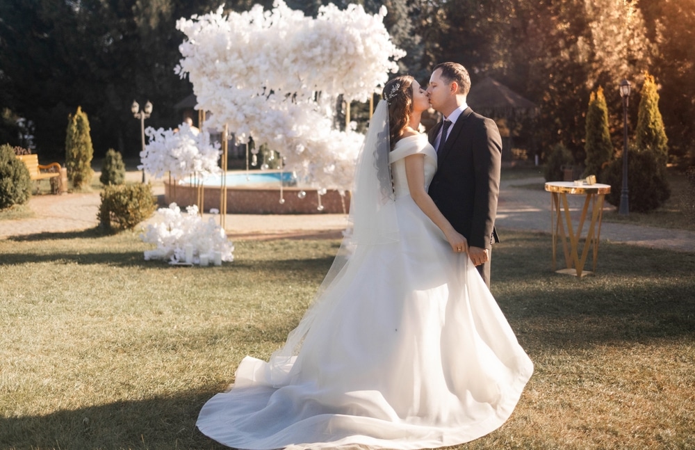 Stylish groom in a suit and a cute brunette bride in a white dress in the forest near a wedding arch decorated with flowers. Wedding portrait of the newlyweds.