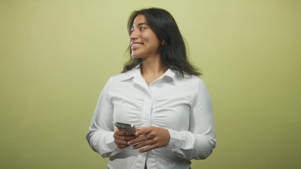 Woman holding smartphone and smiling while looking down in studio, wearing white shirt and relaxed posture; confidence.