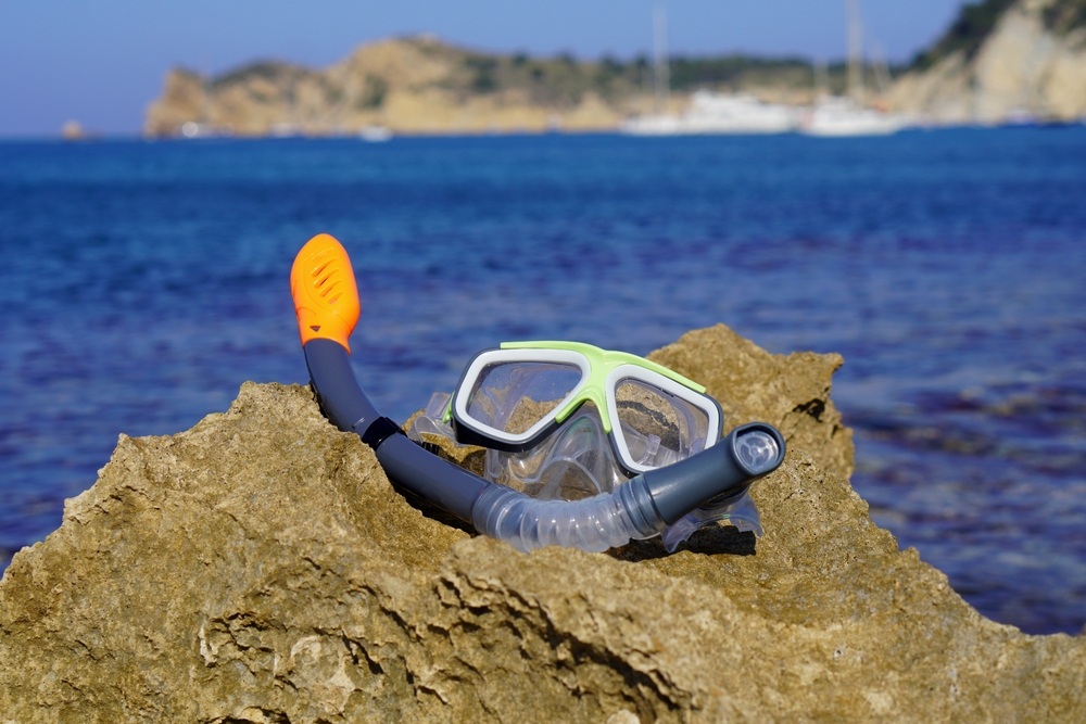 Snorkel and goggles rest on a rock in a sunny Javea cove, with boats floating in the Mediterranean background.