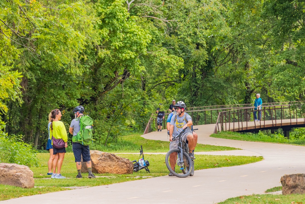 Arkansas, AUG 10 2024 - Cyclists and hikers gather on a winding paved trail surrounded by dense green foliage and a wooden bridge in the distance.